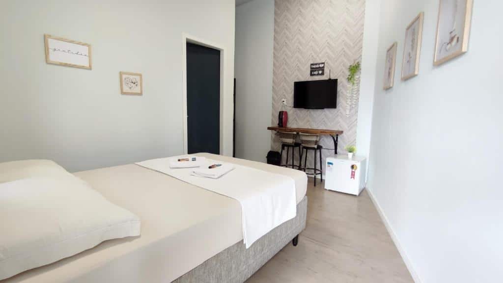 Suíte da Park House Pousada, de 12 m², com uma cama de casal, com lençol branco, travesseiro e duas toalhas de banho brancas dobradas em cima. À frente, há um frigobar, um balcão com duas banquetas e uma TV.