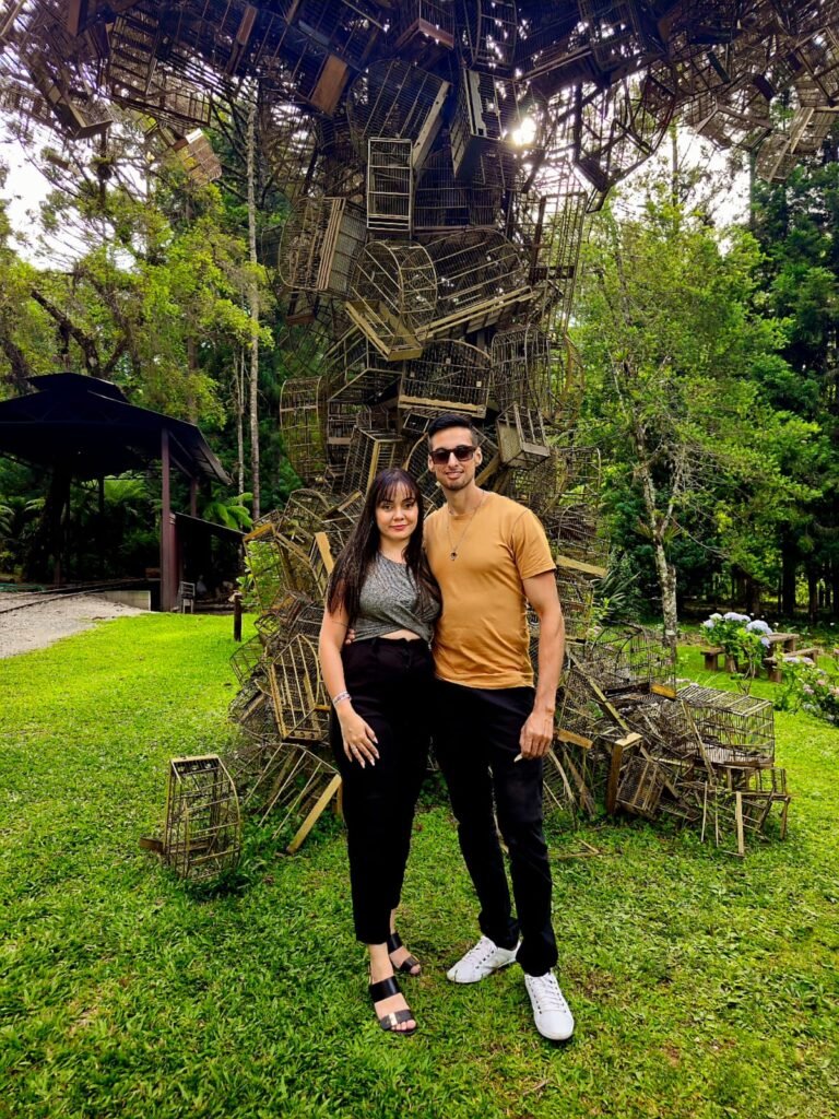 Um homem e uma mulher estão sorrindo em frente a uma grande escultura ao ar livre feita de gaiolas de pássaros empilhadas, cercadas por árvores e grama.