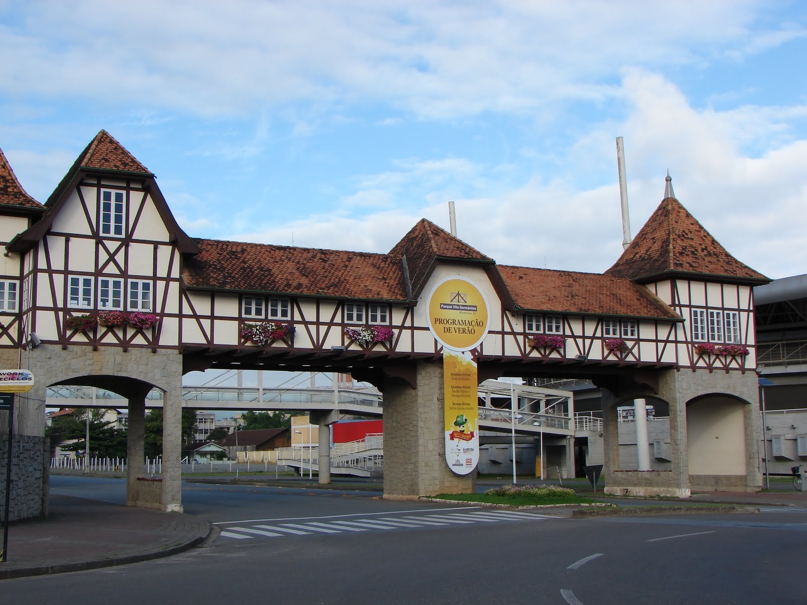 Parque da Vila Germânica em estilo de ponte, com estrutura de madeira e telhados vermelhos, que se estende sobre uma estrada; apresenta uma placa de evento e floreiras sob as janelas. Imagem para ilustrar post sobre o que fazer em Blumenau.