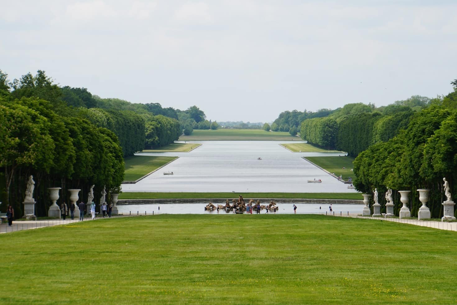 Um longo canal retangular cercado por árvores e estátuas em um jardim formal, com pessoas caminhando por perto. É no Parque de Versalhes.