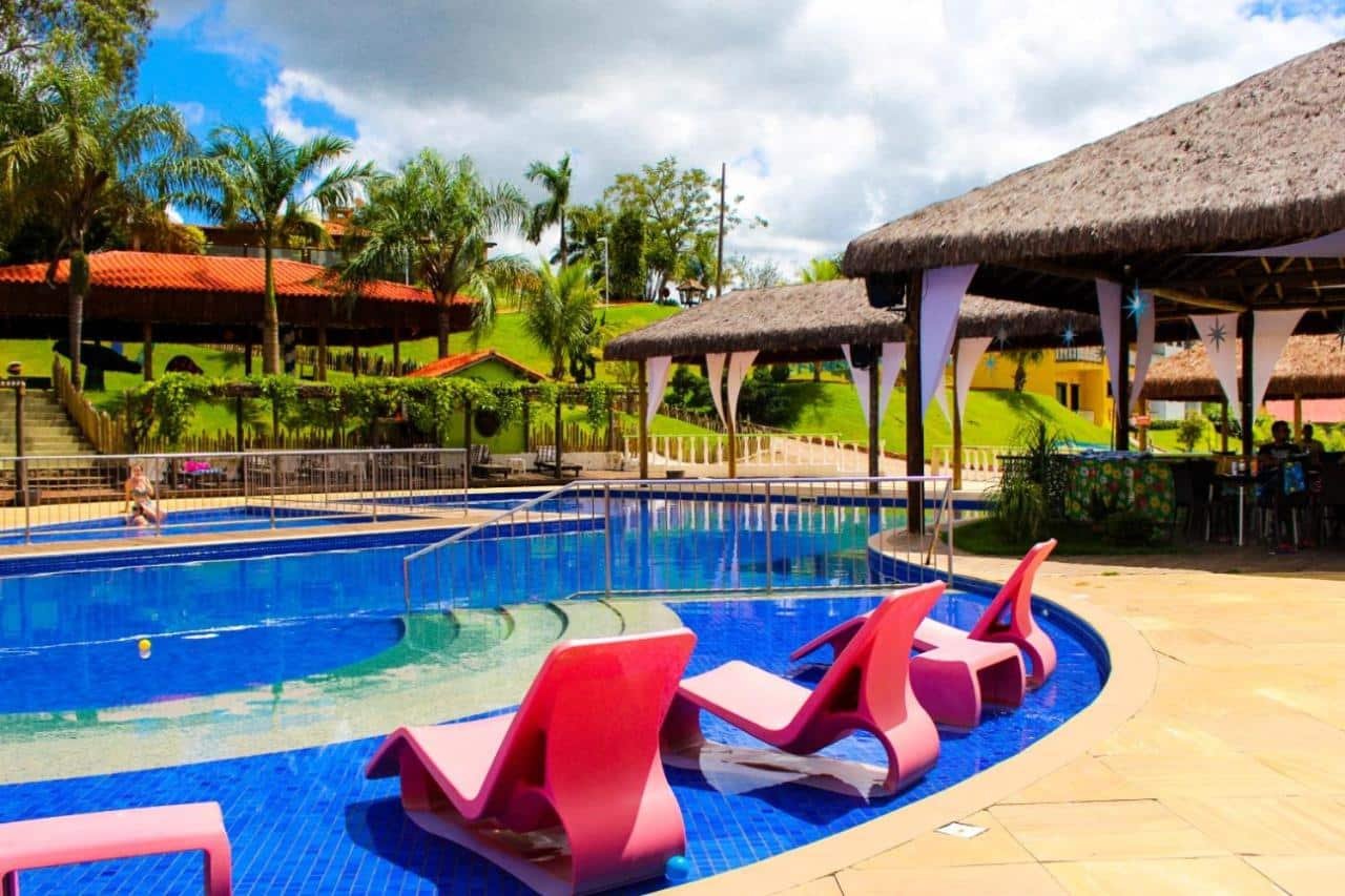A imagem mostra uma área de lazer ao ar livre com uma piscina cercada por espreguiçadeiras cor-de-rosa. Ao redor da piscina, há quiosques com telhados de palha e colunas decoradas com tecidos brancos, criando uma atmosfera tropical. Ao fundo, é possível ver um gramado verde com palmeiras e uma colina suave, além de um céu parcialmente nublado. Algumas construções com telhados vermelhos estão espalhadas pela área, sugerindo uma estrutura de resort ou hotel. O ambiente transmite tranquilidade e lazer em meio à natureza. Esse é o Parque do Avestruz Eco Resort, um dos hotéis-fazenda em Minas Gerais.