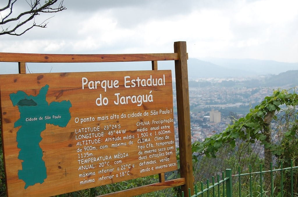 Placa de madeira do Parque Estadual do Jaraguá com detalhes geográficos, com vista para uma paisagem urbana com céu nublado ao fundo.