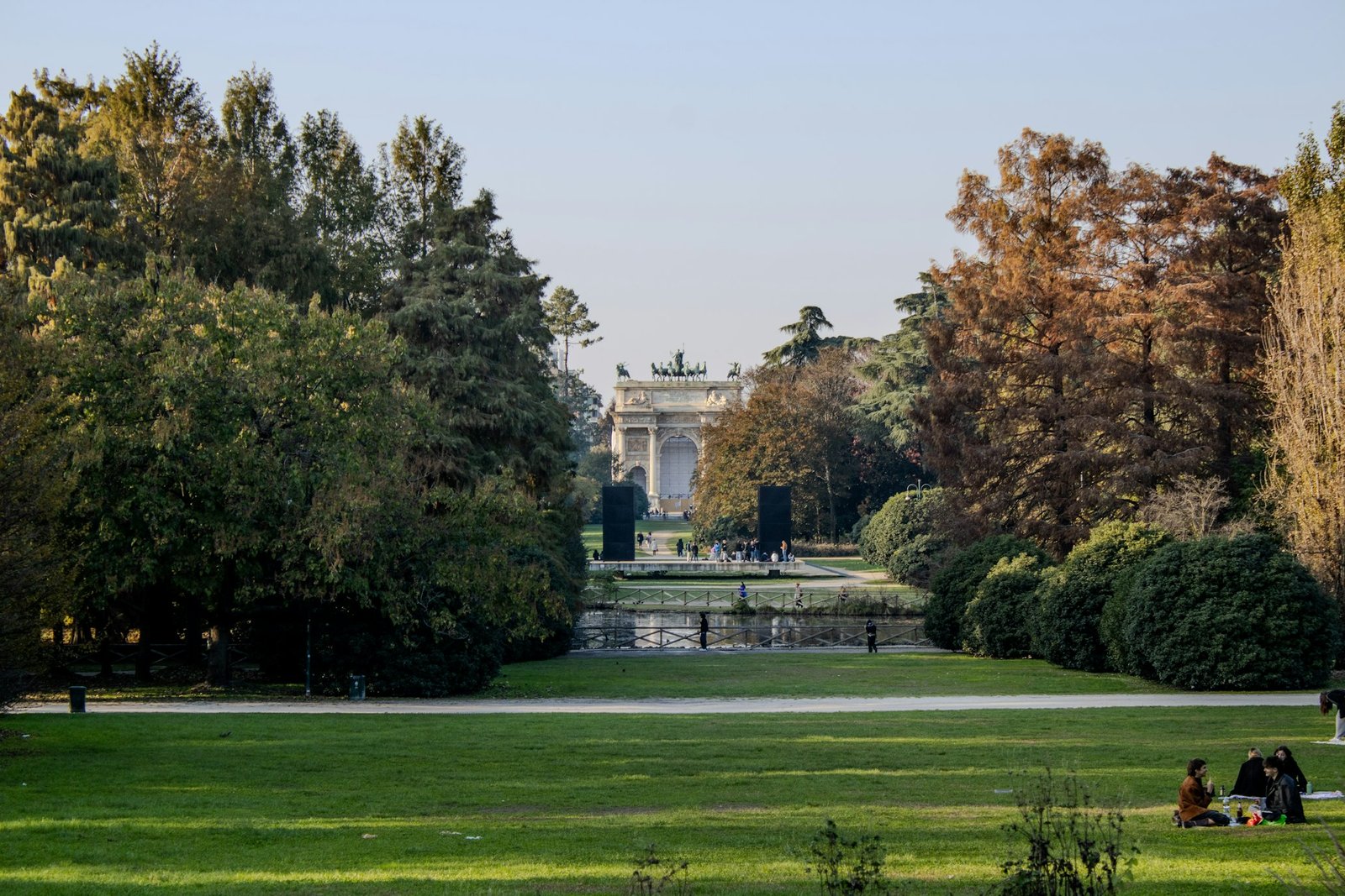 Parque Sempione com árvores espalhadas e pessoas sentadas na grama. O Arco da Paz é visível à distância, atrás de um lago, emoldurado por árvores.