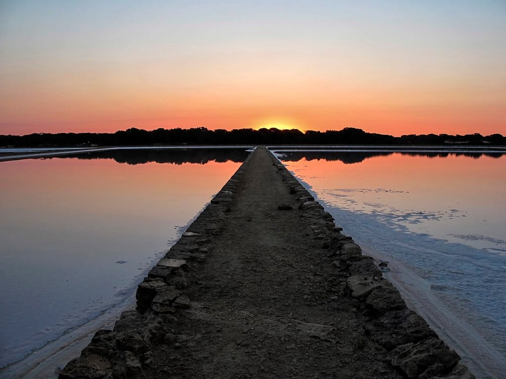 Um caminho estreito ladeado de pedras se estende até o horizonte, ladeado por águas calmas e reflexivas sob um céu de pôr do sol no Parque Natural de Ses Salines.