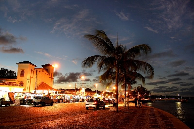 A Passarela do Álcool em Porto Seguro. Está de noite e a rua está iluminada perto da praia. Há coqueiros, carros passando e várias barraquinhas iluminadas.