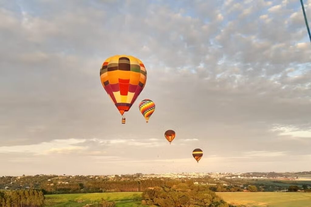 Quatro balões de ar quente coloridos flutuam em um céu parcialmente nublado acima de uma paisagem de Boituva.