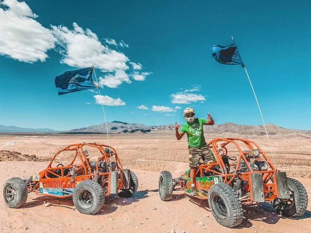 A foto mostra um dois buggys no deserto, um ao lado do outro. Em um deles estã um homem em pé e de capacete.