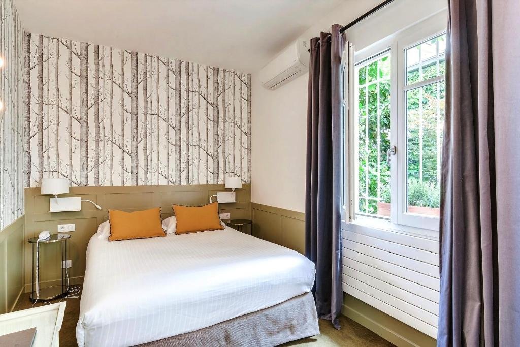 A imagem mostra um quarto acolhedor com uma cama de casal grande e lençóis brancos no Passy Eiffel, um dos hotéis com café da manhã em Paris. A parede atrás da cama tem um papel de parede com um padrão de árvores em tons de cinza e branco. Há duas almofadas decorativas laranja sobre a cama. Ao lado da cama, há mesas de cabeceira com luminárias modernas fixadas na parede. Uma janela grande com cortinas cinza permite a entrada de luz natural e oferece uma vista para o exterior com vegetação. O ambiente é tranquilo e bem decorado, criando uma atmosfera confortável.
