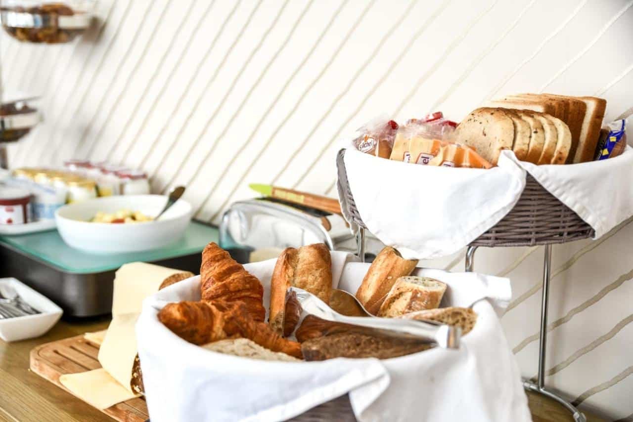 A imagem mostra uma mesa de café da manhã com várias opções de pães e acompanhamentos no Passy Eiffel. Em primeiro plano, há cestas com croissants, baguetes e pães de forma. Ao fundo, há tigelas com manteiga, geleias e uma salada de frutas. Também são visíveis pacotes individuais de biscoitos e uma torradeira. A parede de fundo tem um padrão de listras diagonais sutis, e a mesa está organizada de forma prática e acolhedora.