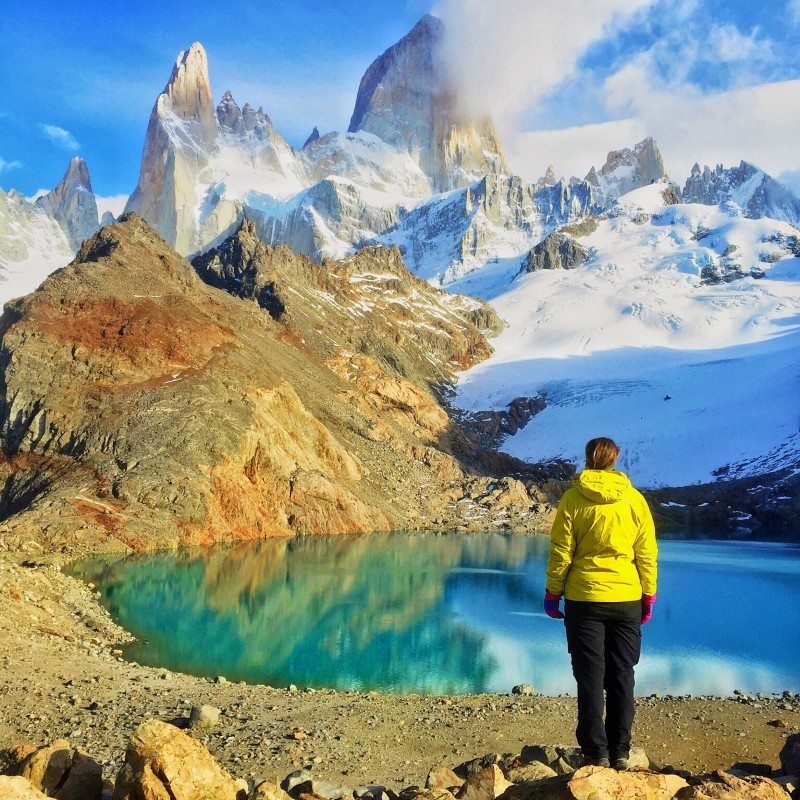 Uma pessoa com uma jaqueta amarela está perto de um lago azul-turquesa, com o Monte Fitz Roy cobertas de neve ao fundo, sob um céu parcialmente nublado.