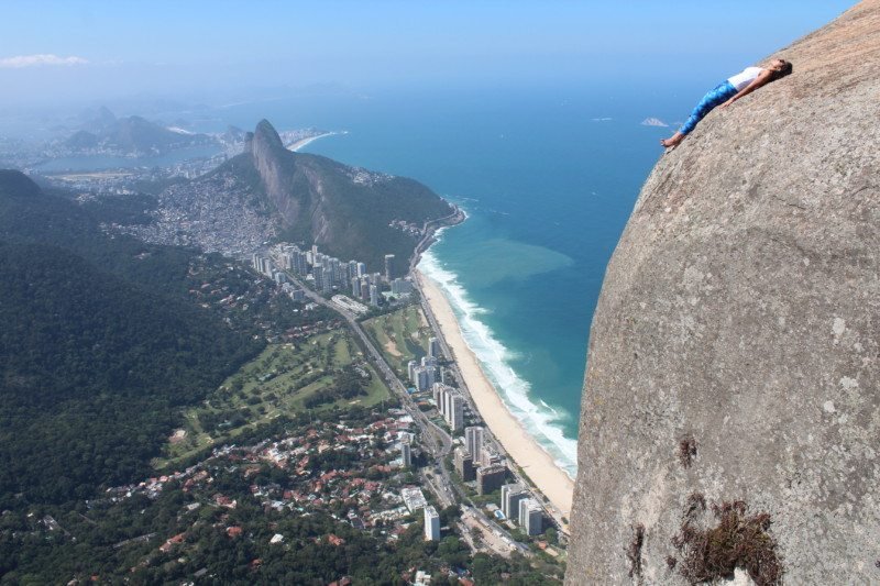 Pessoa deitada em uma grande rocha com vista para uma paisagem urbana costeira com praia, oceano e terreno montanhoso ao fundo. Essa é a paisagem de uma das trilhas no Rio de Janeiro.