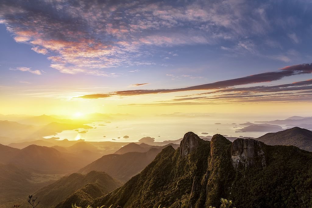 Nascer do sol sobre uma paisagem montanhosa com uma mistura de nuvens e céu limpo. A vista dá para uma baía pontilhada de pequenas ilhas. Essa é a Pedra da Macela, acessada por uma das trilhas em SP.