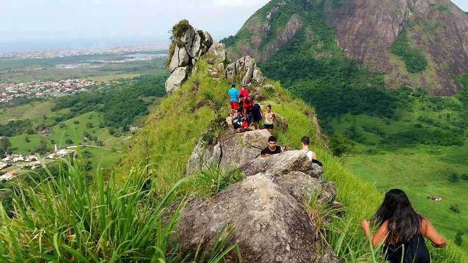 Pessoas caminhando em uma crista rochosa estreita cercada por uma vegetação verdejante, com uma cidade e uma grande colina ao fundo.