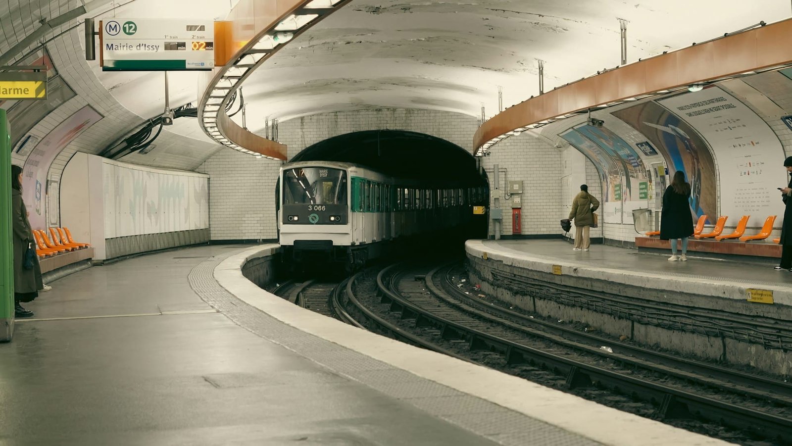 Um trem do metrô está se aproximando de uma plataforma subterrânea curva. Duas pessoas esperam do lado direito. A estação tem assentos laranja.