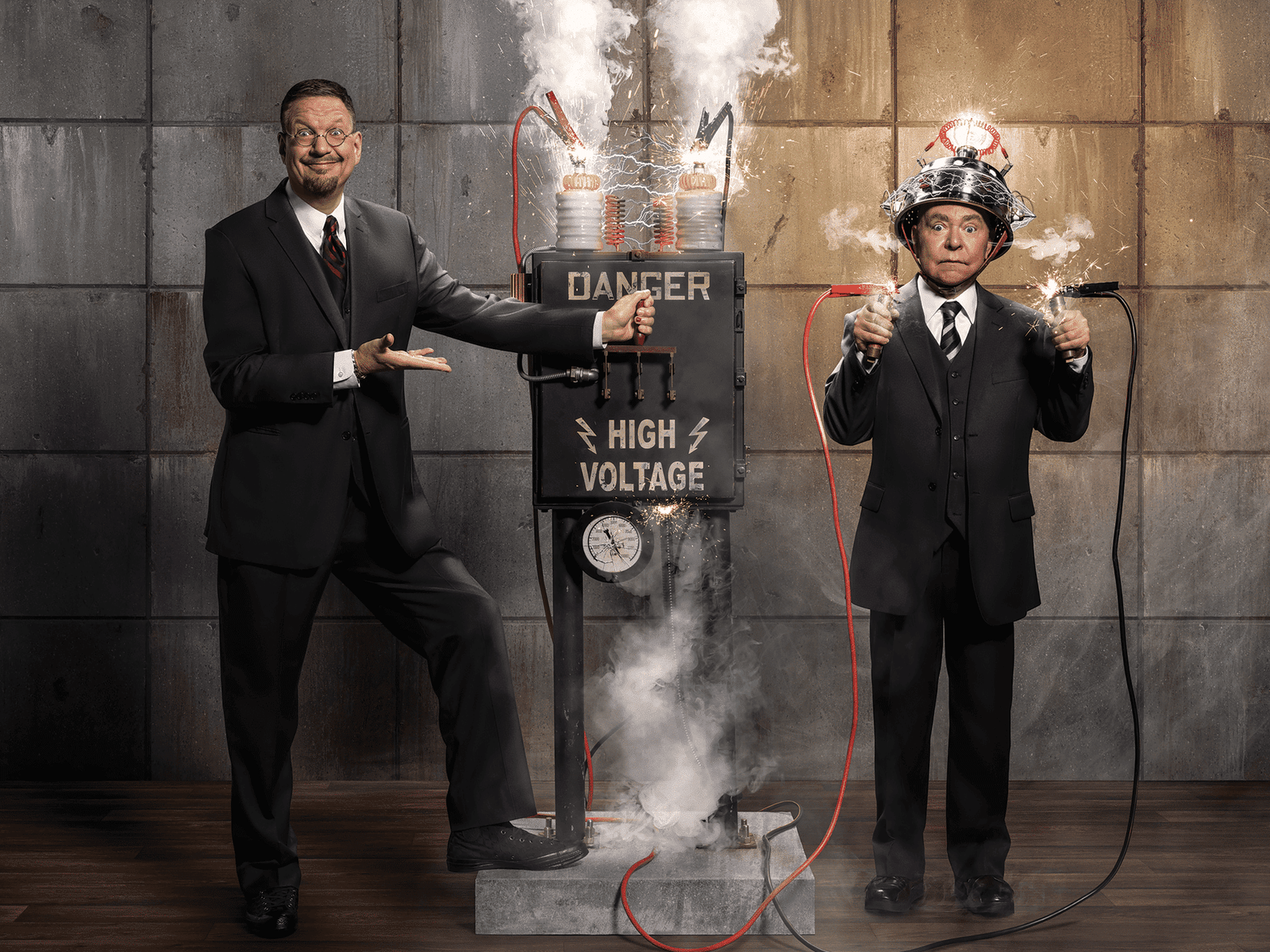 Penn & Teller de terno estão de pé ao lado de uma máquina de alta voltagem com lâmpadas acesas. Um aponta para ela; o outro usa um capacete com fios, segurando cabos.