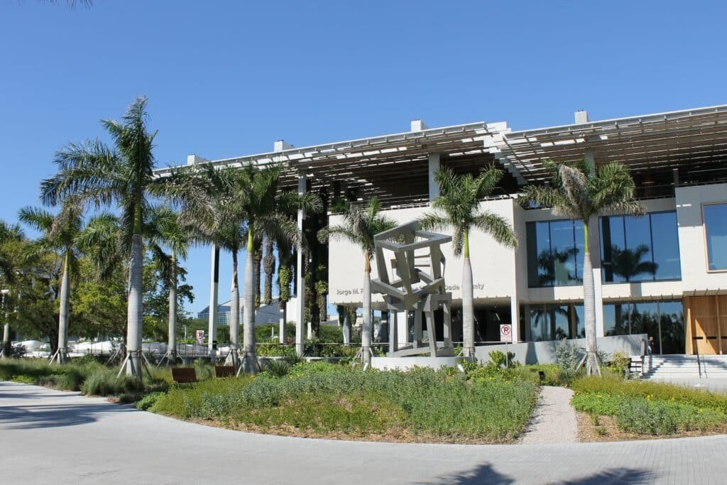 Entrada do Pérez Art Museum Miami (PAMM). Em frente há um jardim, algumas árvores e uma escultura.