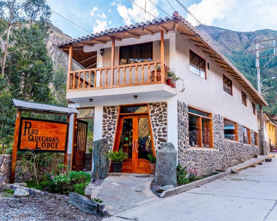 A imagem retrata uma casa de dois andares chamada “Peru Quechuas Lodge”, situada em um ambiente montanhoso. A parte inferior da casa é construída com pedras, enquanto a parte superior é de cor creme, com uma varanda de madeira. O letreiro à esquerda da entrada principal exibe o nome da pousada. O cenário natural ao redor da casa é composto por vegetação e montanhas, sob um céu claro.