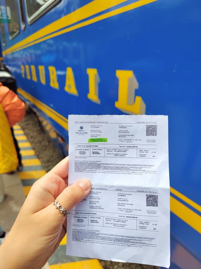 bilhete do Perurail sendo segurado por uma mão feminina esquerda, usando um anel de estrelas no polegar. Atrás da mão é possível enxergar um vagão de trem azul