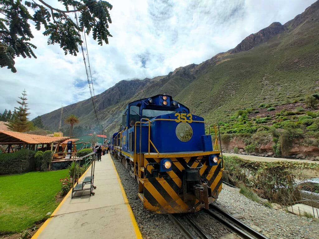 visão panorâmica do trem da Perurail vista de frente para o vagão condutor, parado na estação. Ao redor é possível enxergar as montanhas peruanas e a plataforma de acesso em ollantaytambo, no vale sagrado dos incas