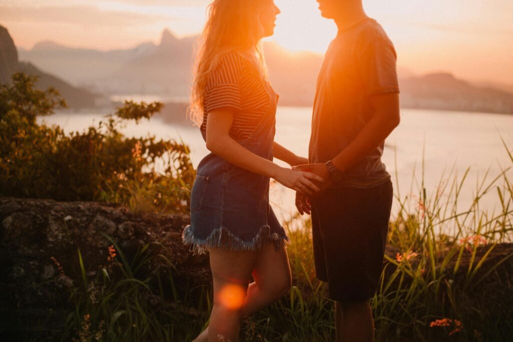 Um casal está perto um do outro no pôr do sol, de frente um para o outro. Eles estão do lado de fora, com uma vista panorâmica e folhagens ao redor.