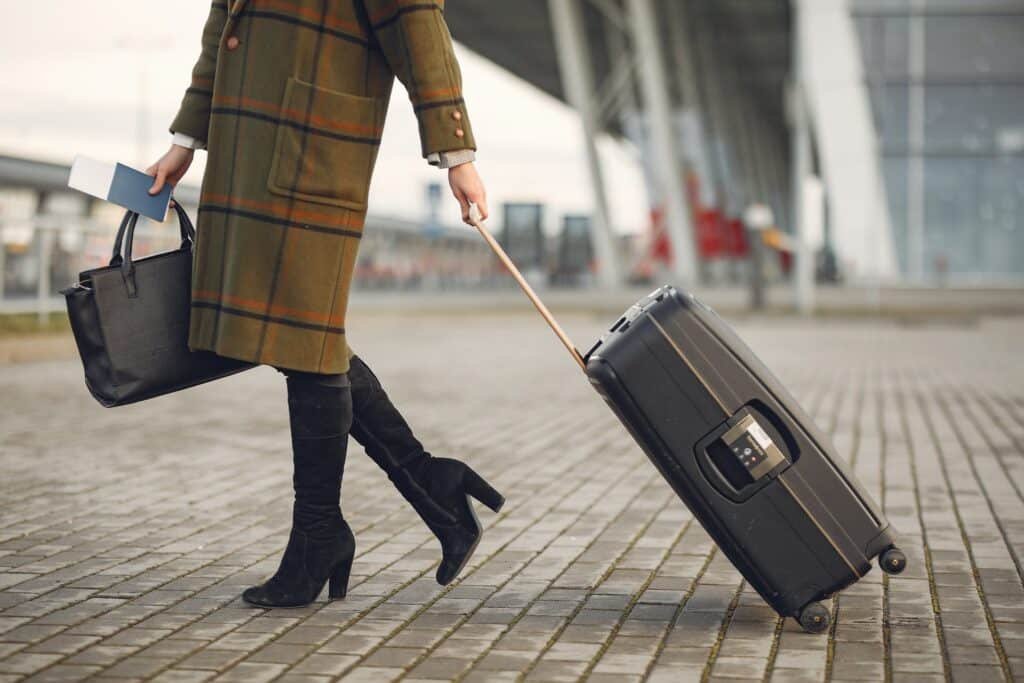 Uma mulher usando um casaco xadrez e botas, ela está puxando uma mala de rodinhas com uma das mãos, com a outra está segurando uma bolsa e um passaporte, para representar seguro bagagem