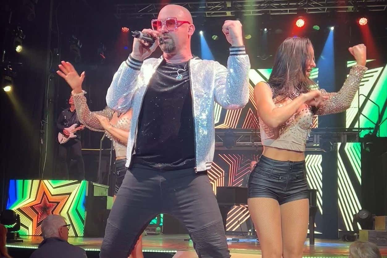 Kyle Martin durante o show Piano Man com uma jaqueta brilhante e óculos de sol rosa canta no palco, ladeado por dançarinos. Luzes coloridas e um cenário são visíveis. A foto ilustra o post de shows em Las Vegas.