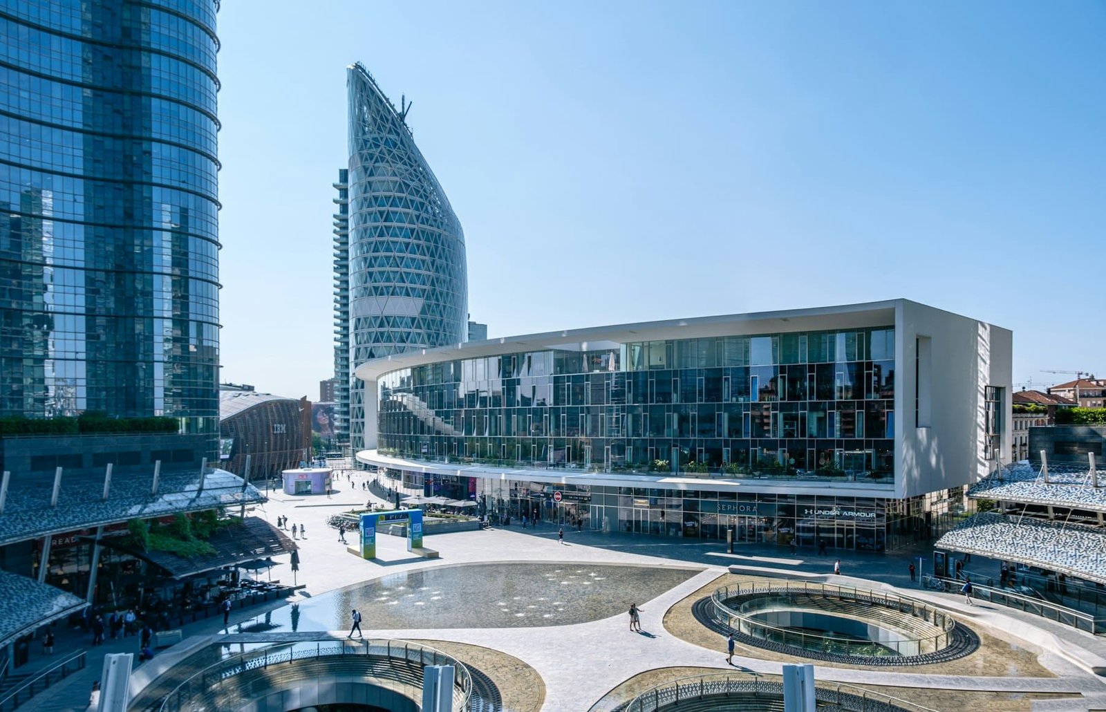 Imagem panorâmica da Piazza Gae Aulenti. Se trata de uma praça urbana moderna com edifícios futuristas, incluindo uma estrutura curva com fachada de vidro e uma torre alta e de formato único. Foto na lista de o que fazer em Milão.