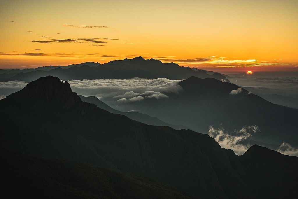 Nascer do sol sobre uma cadeia de montanhas com picos recortados, nuvens abaixo e um céu laranja brilhante iluminando o horizonte. Esse é o Pico dos Marins, acessado por uma das trilhas em SP.