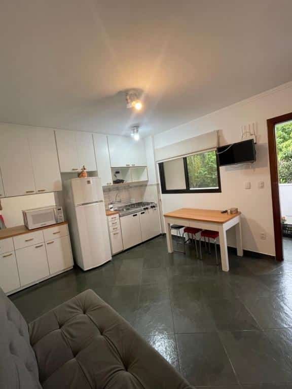 Cozinha e sala bem espaçosas em Piemonte Flat Serra Neg SP. No canto inferior esquerdo vemos parte do sofá, e ao fundo há uma cozinha planejada com armários brancos, pia, geladeira, micro-ondas e mesa com três bancos. No topo da parede que está a mesa há uma TV e uma janela lado a lado.