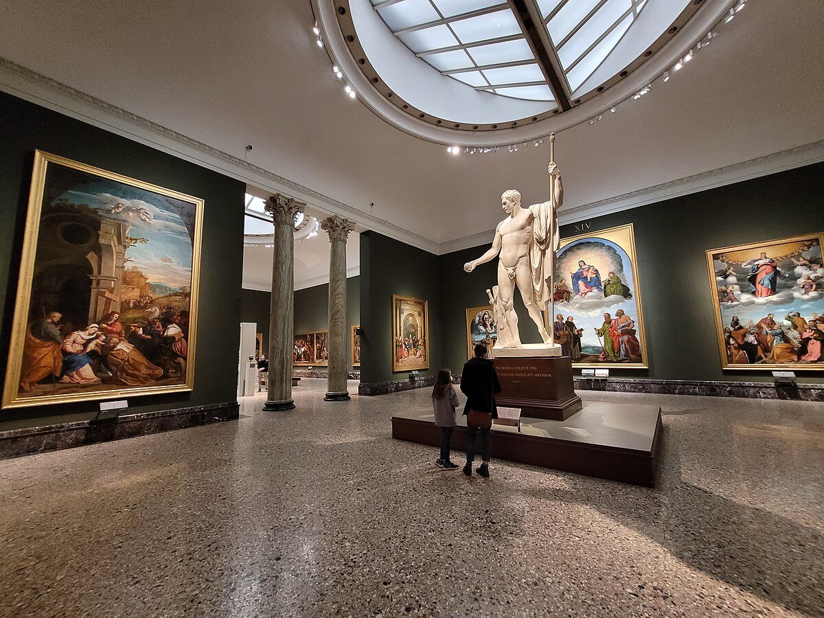 Uma das salas na Pinacoteca di Brera com grandes pinturas clássicas e uma estátua central. Duas pessoas estão de pé observando a obra de arte.