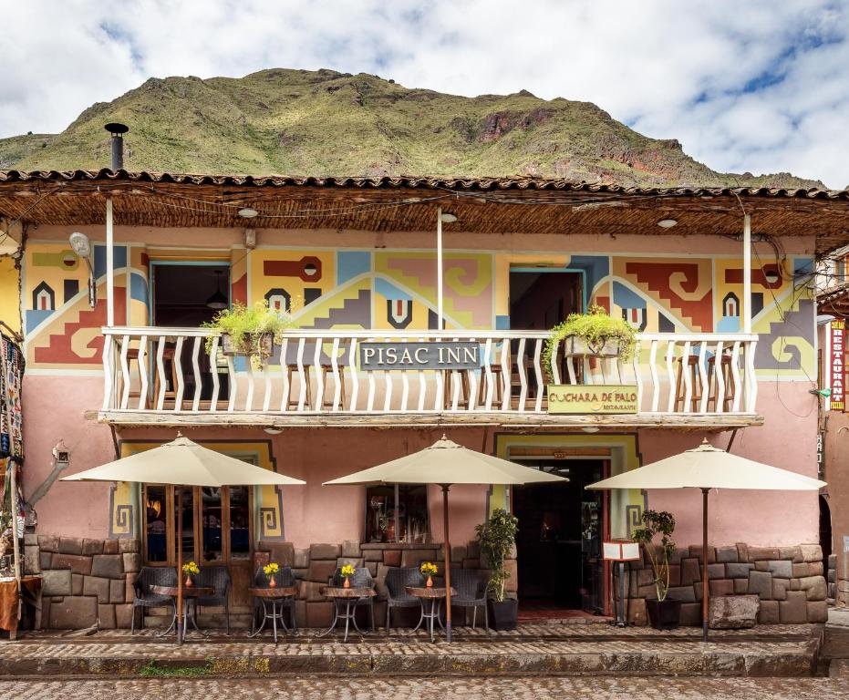 vista frontal do Pisac Inn, onde há uma construção de dois andares, pintada de rosa clara e com uma varanda de madeira no segundo andar. Na calçada, em frente a entrada, há várias mesas e cadeiras, acompanhadas de guarda-sóis.