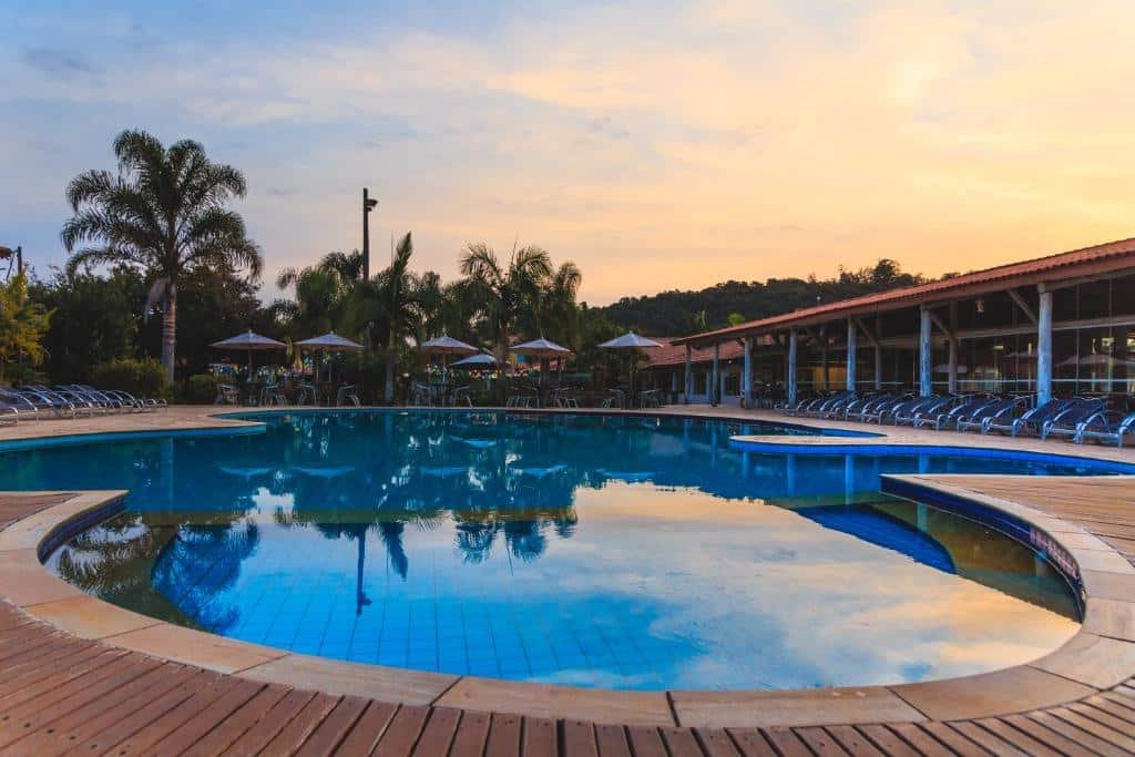 Uma piscina redonda ao ar livre do Hotel Fazenda Hípica Atibaia, um dos hotéis fazenda em São Paulo, cercada por espreguiçadeiras e palmeiras ao pôr do sol, com um edifício ao fundo.