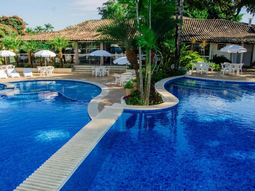 Área de piscina externa no Hotel Recanto da Cachoeira, um dos hotéis fazenda em São Paulo, com formas curvas, cercada por espreguiçadeiras e guarda-sóis. Uma ponte atravessa a piscina; um prédio está ao fundo.