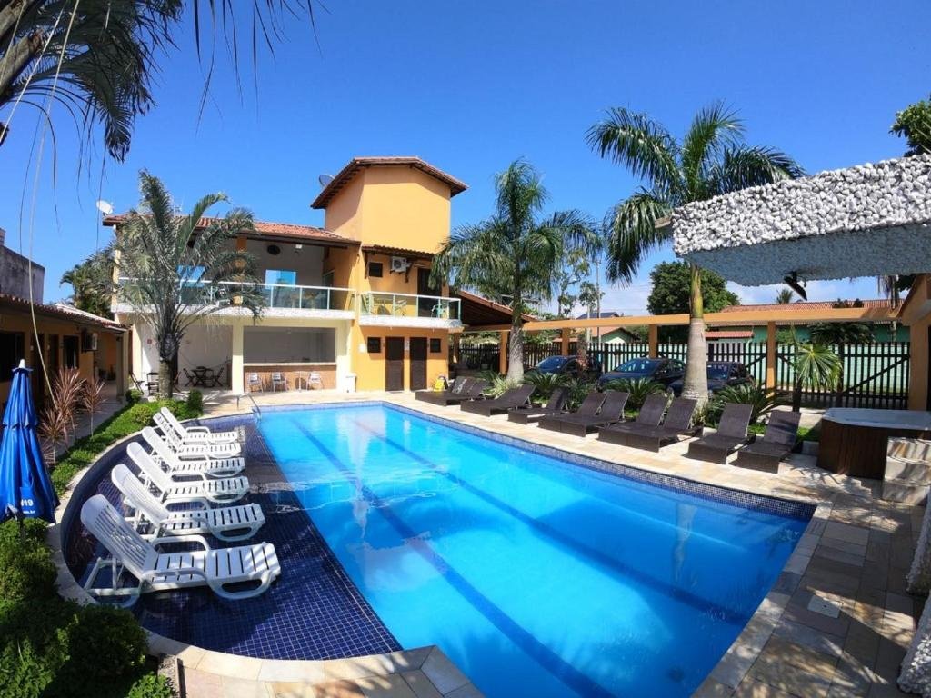 Área de piscina ao ar livre com espreguiçadeiras e palmeiras. Um prédio amarelo de dois andares está ao fundo sob um céu azul claro na Pousada Acqua Bella.