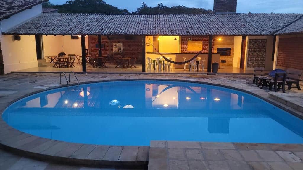 Área externa de uma casa para aluguel de temporada com piscina, área para churrasco, mesas, cadeiras e uma rede.