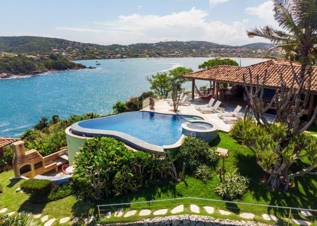 Vista de cima da Cliffside Luxury Inn com piscina no centro em volta de uma jardim e do lado direito da imagem a estadia de frente com a piscina vista do mar. Representa airbnb na Barra da Tijuca.