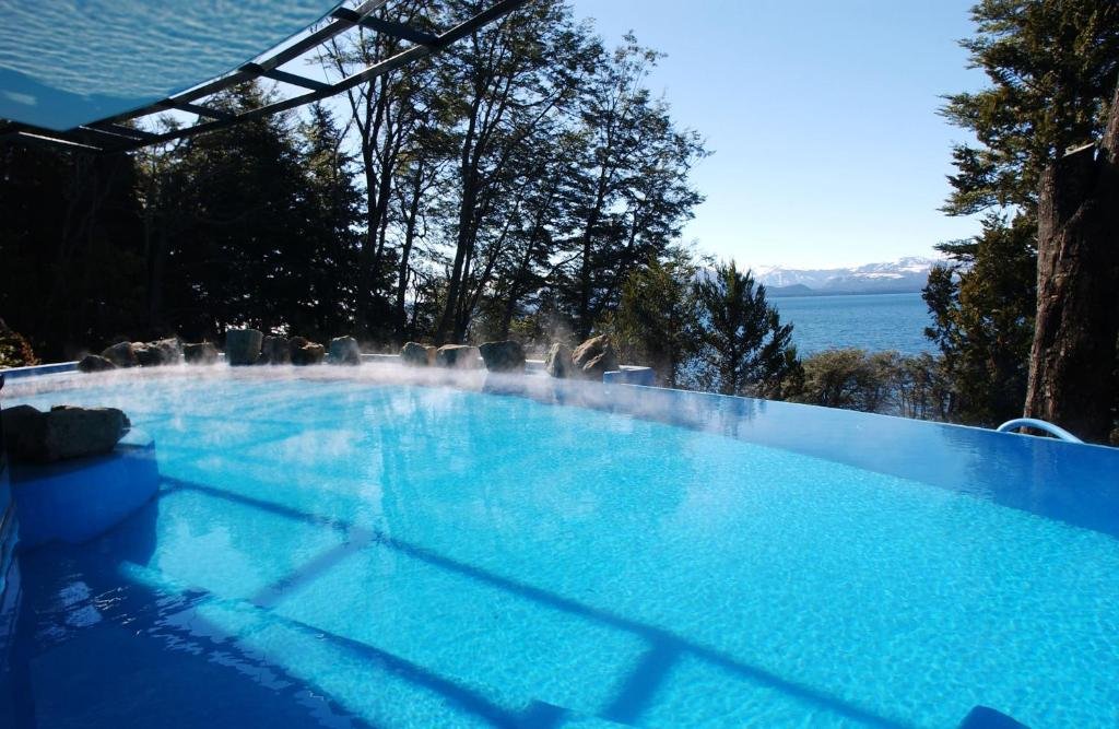 Piscina ao ar livre do Design Suites Bariloche com vista para árvores e ao fundo para o rio. Representa hotéis em Bariloche