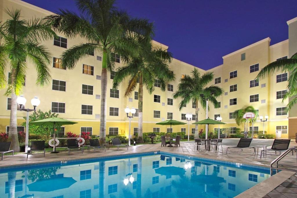 Vista noturna de um pátio de hotel com uma piscina iluminada, palmeiras, espreguiçadeiras e uma cesta de basquete.