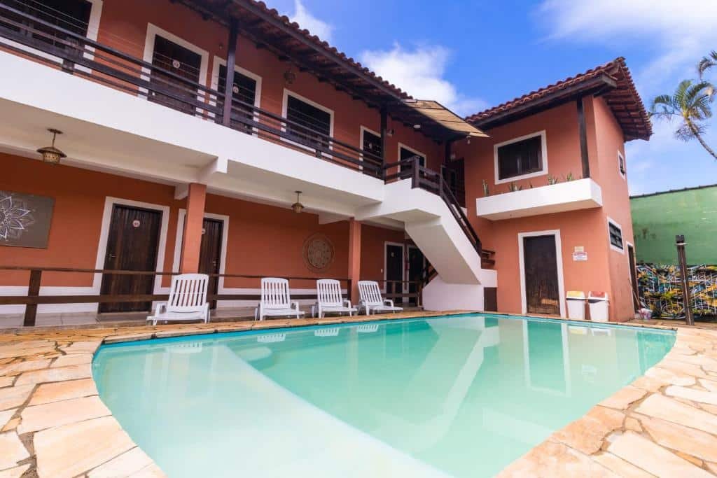 Parte externa da HOTELARE Bóra Morá Pousada, uma piscina com algumas espreguiçadeiras ao redor, a construção possui varandas e é pintada em tons de vermelho escuro