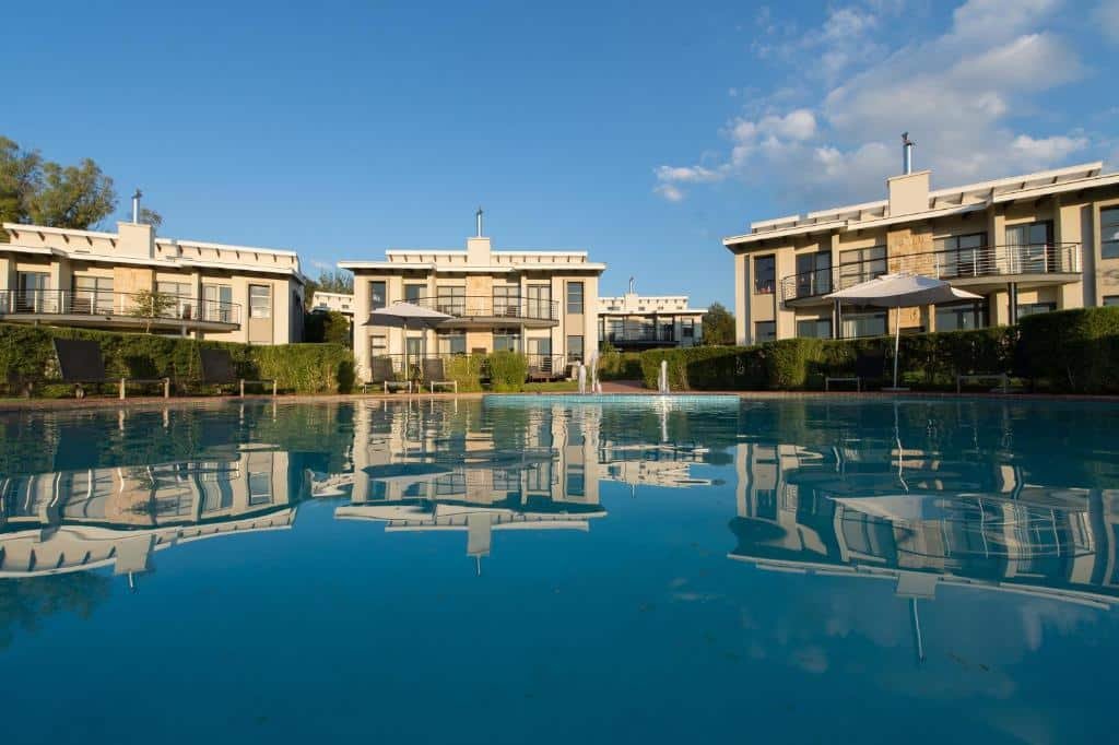 Moradias modernas do The Fairway Hotel, Spa & Golf Resort
com janelas grandes são refletidas em uma piscina azul-clara. Guarda-sóis e vegetação estão ao fundo. Representa Joanesburgo.
