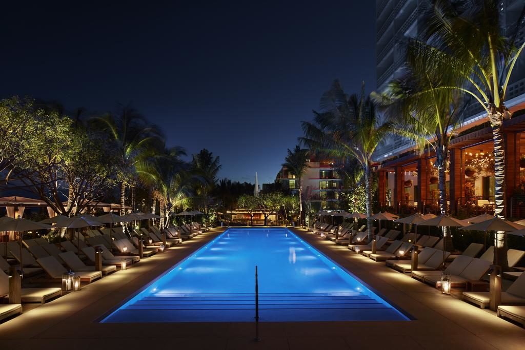A imagem mostra uma piscina iluminada à noite no The Miami Beach EDITION, um dos hotéis em Miami Beach recomendados. A piscina é retangular, com luzes azuis submersas que destacam a água. Ao redor da piscina, há espreguiçadeiras alinhadas sob guarda-sóis brancos e árvores iluminadas, criando um ambiente acolhedor. À direita, um edifício com varandas e luzes acesas sugere um hotel ou resort. Lanternas decorativas estão dispostas ao longo das espreguiçadeiras, aumentando o charme e a atmosfera relaxante do local.