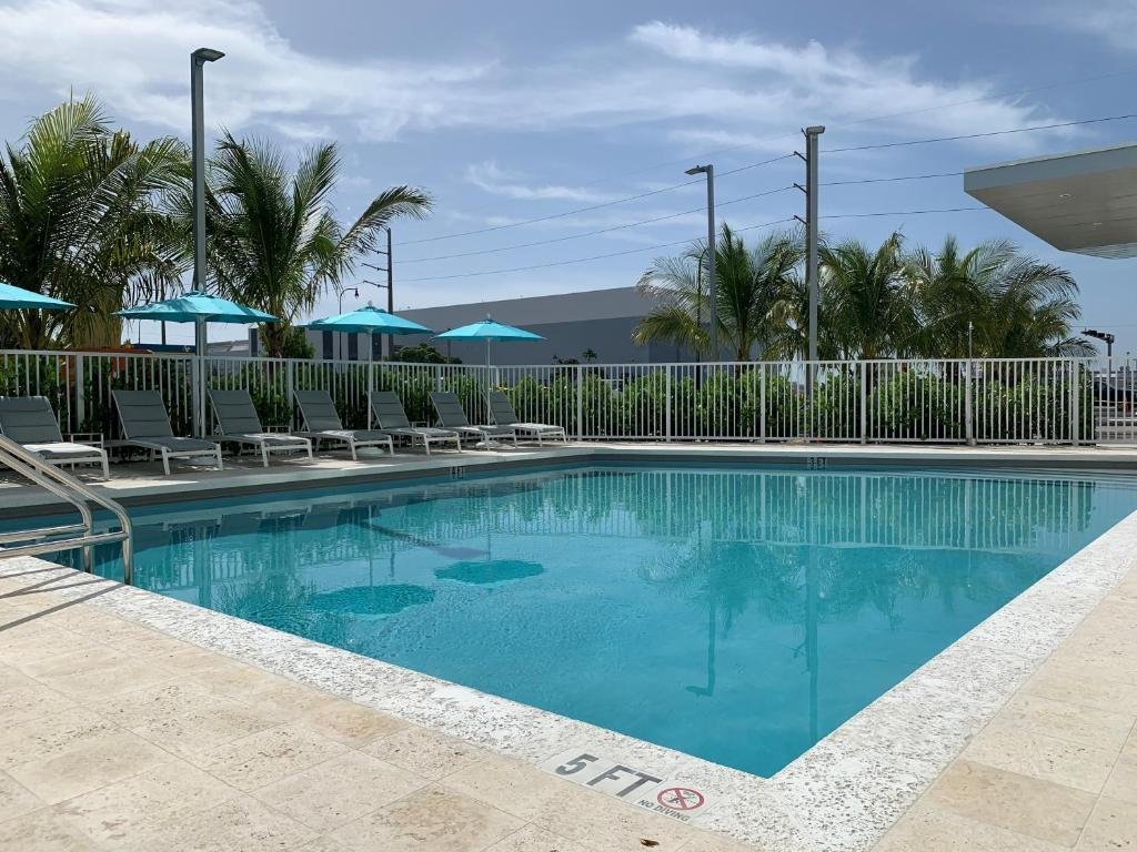 Uma piscina ao ar livre com água azul clara, cercada por espreguiçadeiras e guarda-sóis azuis. Palmeiras ao fundo.
