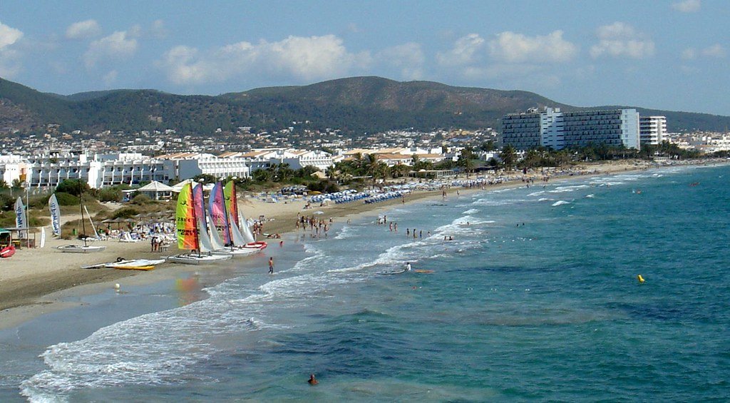 Uma praia de areia com veleiros coloridos, pessoas tomando sol e uma vista distante de edifícios tendo como pano de fundo colinas. Essa é a Playa d