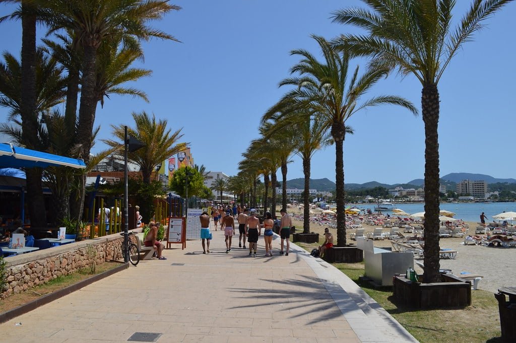 Pessoas caminhando em um calçadão cercado por palmeiras, perto de uma praia de areia com banhistas e guarda-sóis, sob um céu limpo. Essa é a Playa de San Antonio, uma das praias em Ibiza recomendadas.