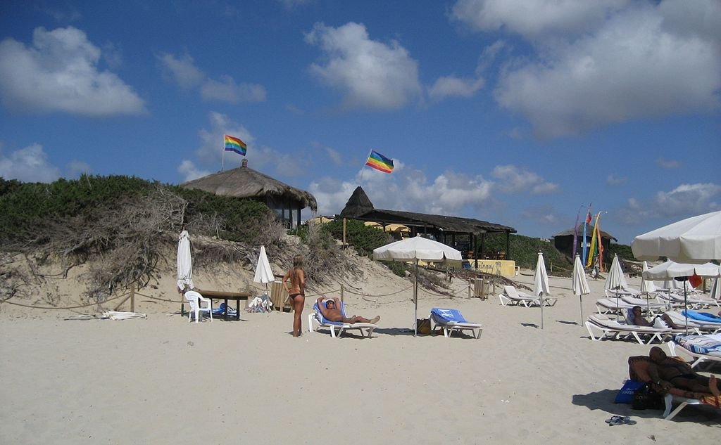 Praia de areia com espreguiçadeiras e guarda-sóis. Duas bandeiras de arco-íris em cabanas ao fundo. Pessoas tomando sol sob um céu azul.