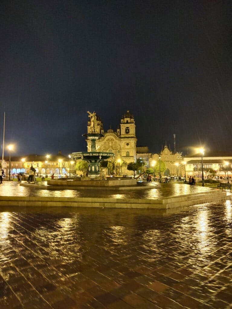 plaza de armas em cusco vista a noite Há várias luzes amarelas acesas e cai uma fina chuva. Bem no centro da imagem há uma fonte com uma estatueta em cima.