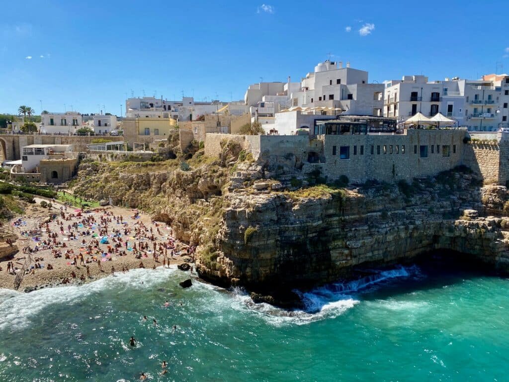 Imagem aérea da pequena praia a esquerda com várias pessoas na faixa de areia e algumas na água azul e verde. Já do lado direito um paredão de pedra com várias casas em cima durante o dia, ilustrando post Hotéis em Puglia.