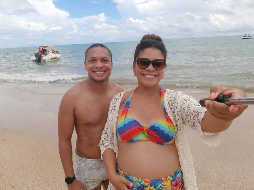 Um casal sorridente tira uma selfie na praia. A mulher segura um bastão de selfie, usando um maiô arco-íris e óculos de sol para representar um dos melhores lugares para lua de mel