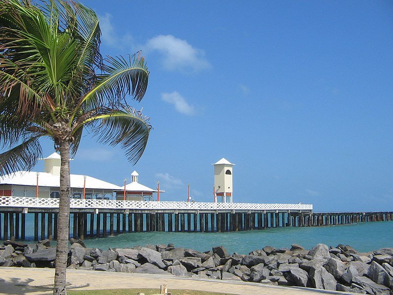 Foto tirada de dia na Praia de Iracema. Na esquerda está a Ponte dos Ingleses, uma das atrações turísticas de Fortaleza. Na areia da praia há várias rochas e pedras. O céu está bem azul e com algumas nuvens. Na esquerda há um coqueiro.