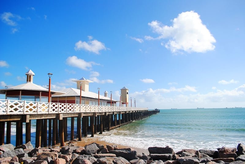 Foto tirada de dia na Praia de Iracema. Na esquerda está a Ponte dos Ingleses, uma das atrações turísticas de Fortaleza. Na areia da praia há várias rochas e pedras. O céu está bem azul e com algumas nuvens.